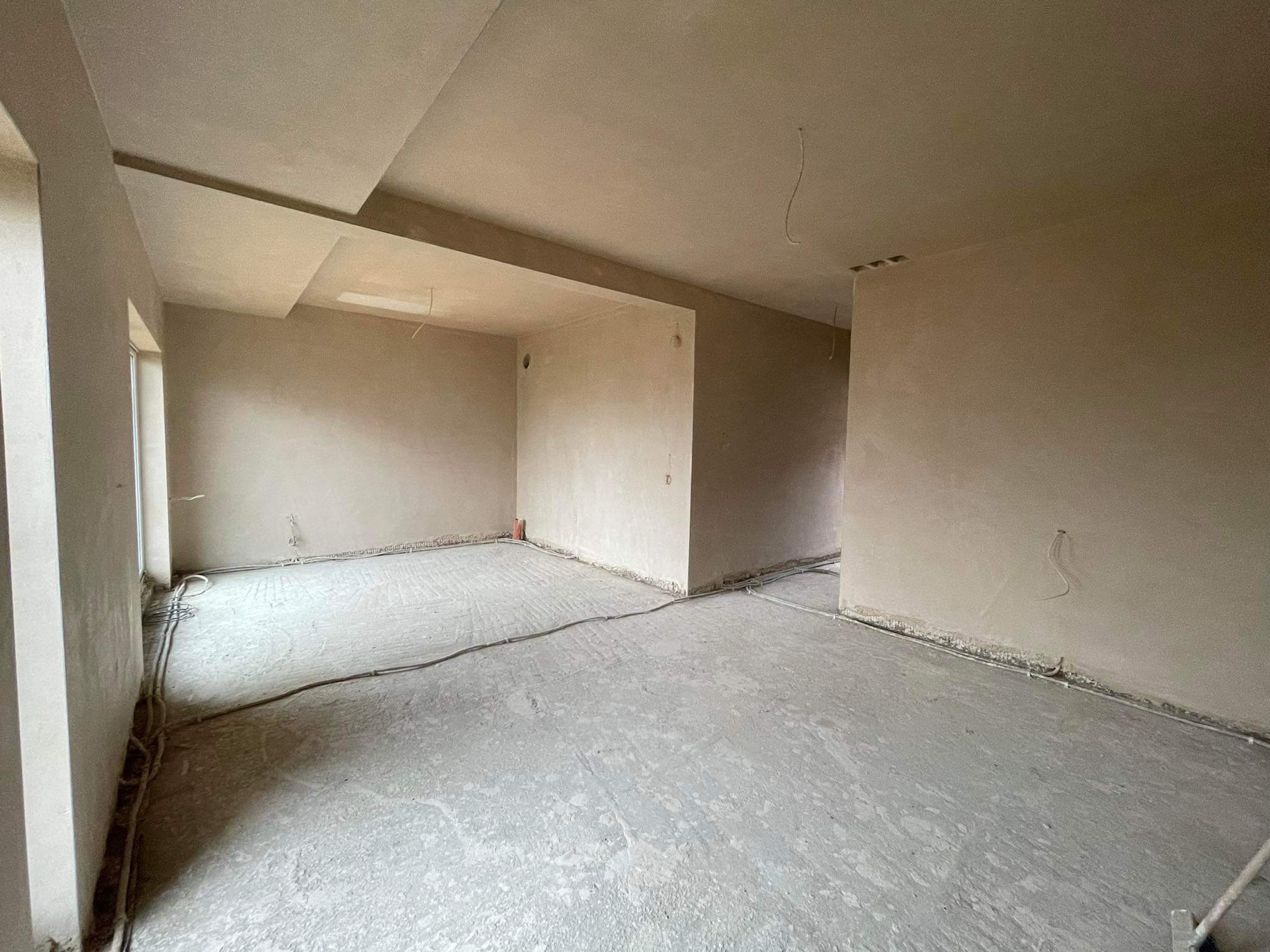 Surowe wnętrze po nałożeniu tynku maszynowego, widoczne kable elektryczne prowadzone po podłodze, sufit z widocznymi łączeniami płyt gipsowych.