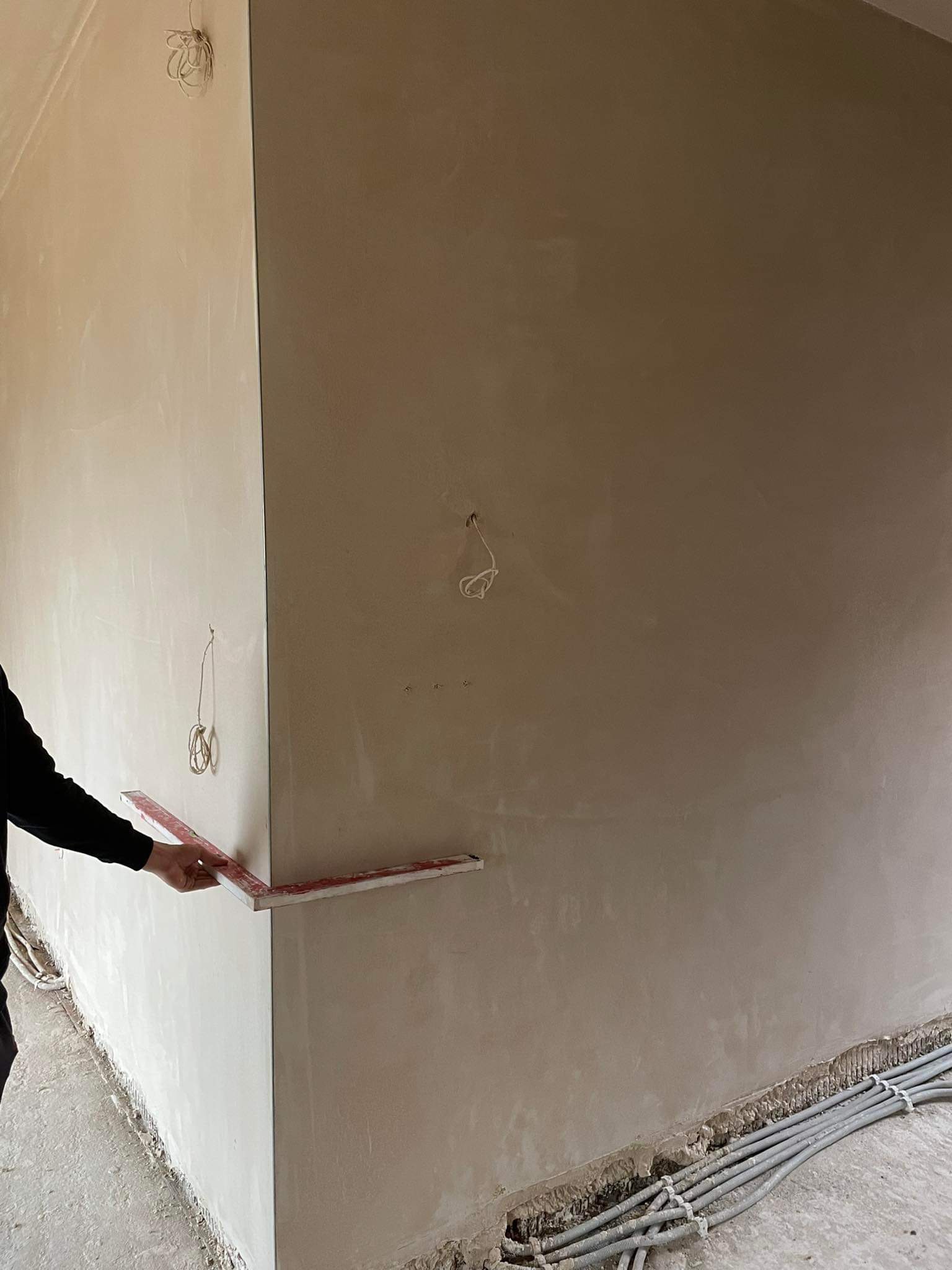 Świeżo otynkowana ściana z wystającymi przewodami elektrycznymi przy podłodze, sprawdzana poziomicą przez pracownika w czarnej bluzie.