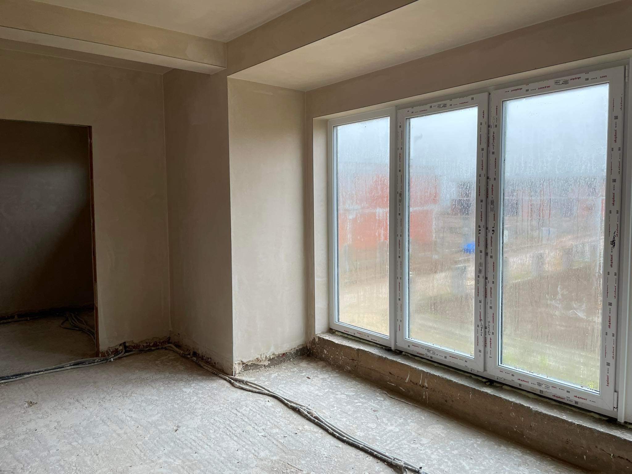 Świeżo otynkowane ściany w pokoju z dużym oknem, widoczne wystające kable elektryczne wzdłuż podłogi, ślady wilgoci na szybach okiennych.