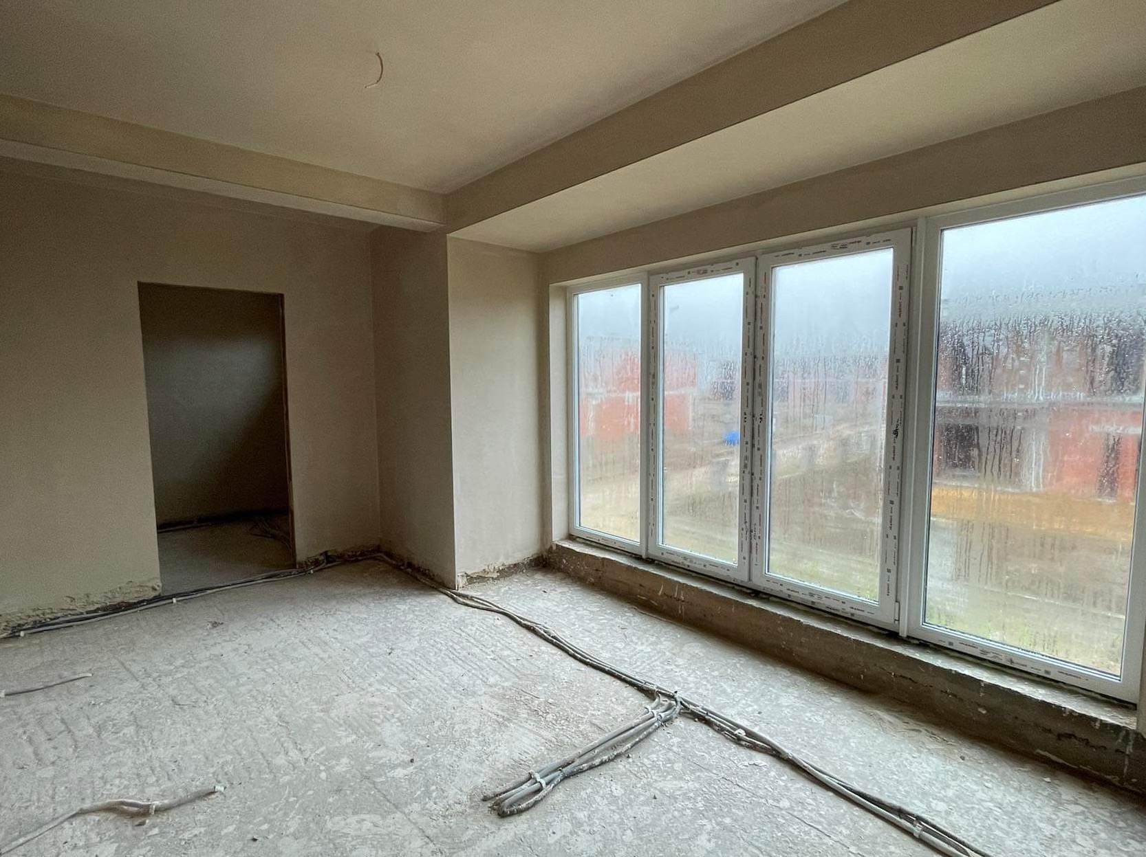 Surowe wnętrze w trakcie prac wykończeniowych: ściany po tynkowaniu maszynowym, widoczne kable elektryczne na podłodze, okno z widokiem na budowę w deszczowy dzień.