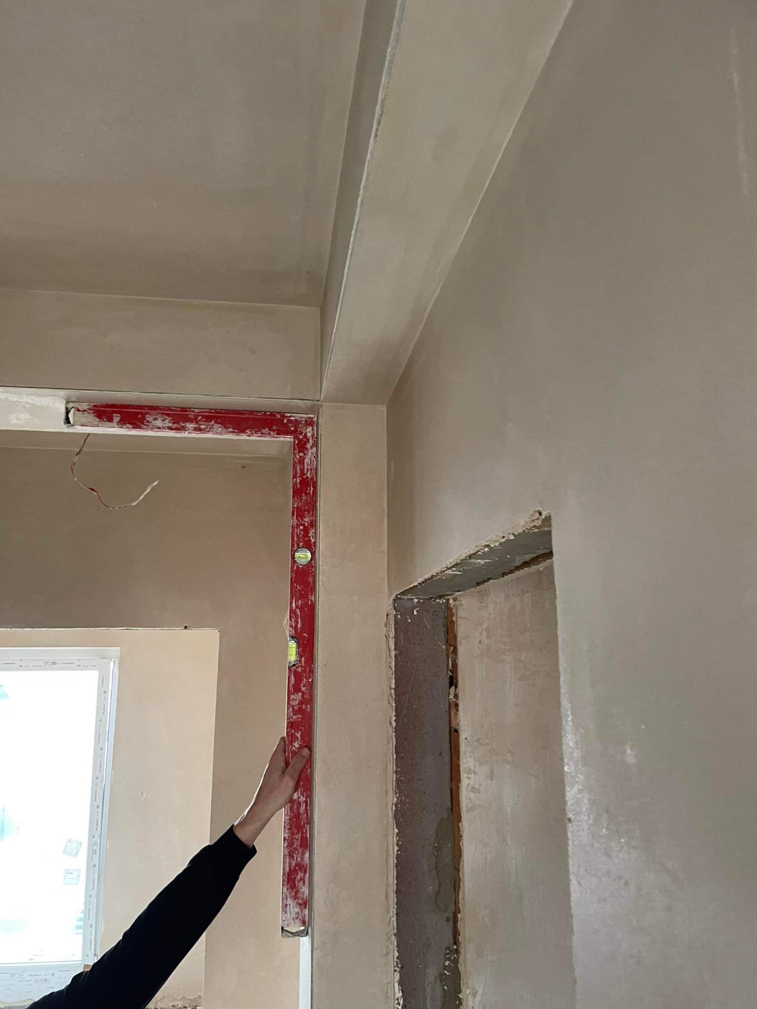 Prace wykończeniowe w Warszawie: Położona świeża warstwa na ścianie w trakcie poziomowania czerwoną poziomicą przy futrynie drzwiowej.