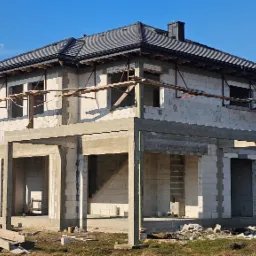 Dwukondygnacyjny dom w stanie surowym otwartym, z widocznymi elementami konstrukcyjnymi i szarym dachem, na tle błękitnego nieba i pola. Widoczne rusztowania i materiały budowlane.