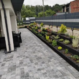 Nowoczesny ogród przydomowy z wielopoziomowymi rabatami z kostki, oświetlony lampami solarnymi, z widokiem na szary płot panelowy i budynek z czerwonej cegły w tle.