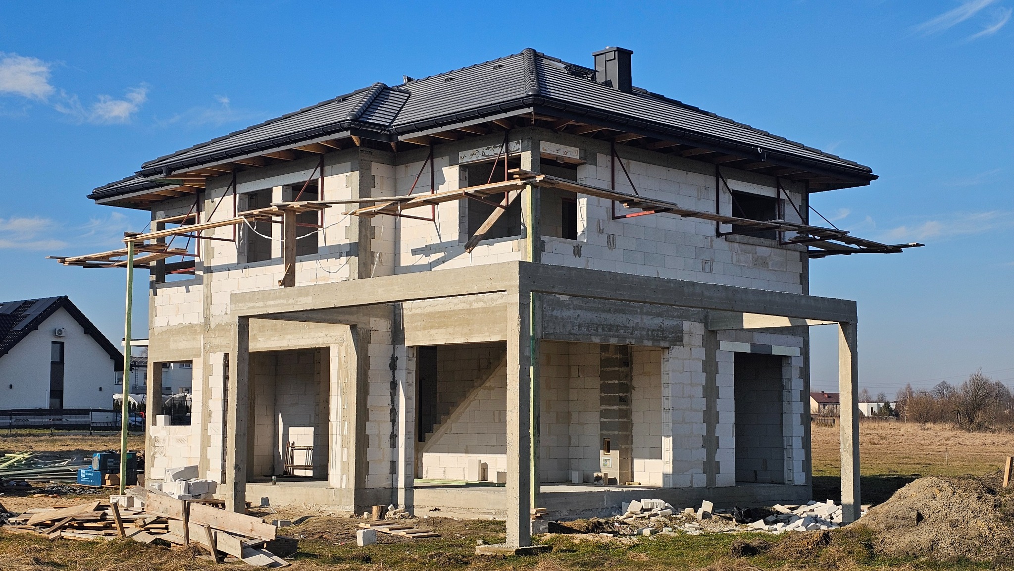 Dwukondygnacyjny dom w stanie surowym otwartym, z widocznymi elementami konstrukcyjnymi i szarym dachem, na tle błękitnego nieba i pola. Widoczne rusztowania i materiały budowlane.