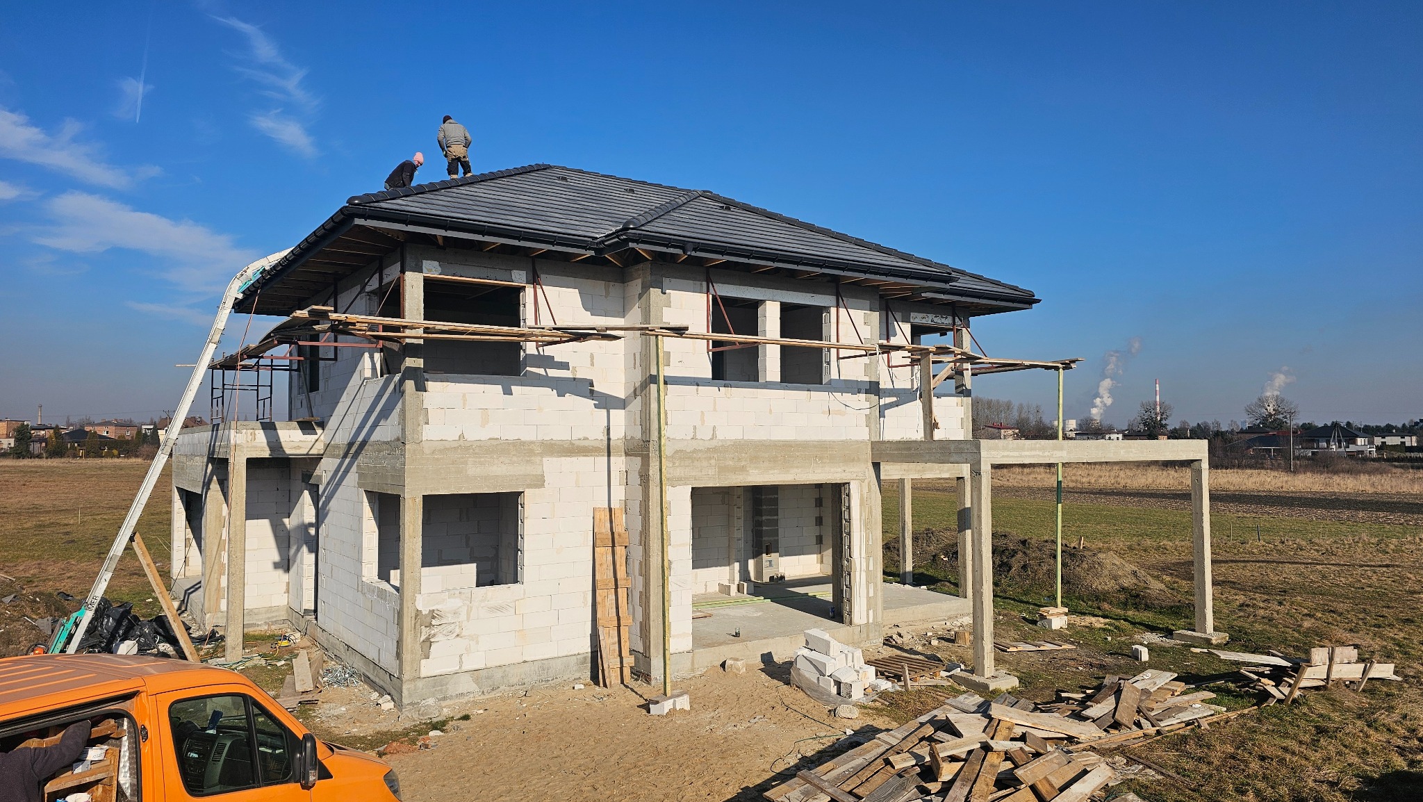 Wznoszenie domu z białych bloczków, częściowo zadaszonego ciemną dachówką. Na dachu dwie osoby. Widoczne rusztowania i materiały budowlane. W tle kominy fabryczne.