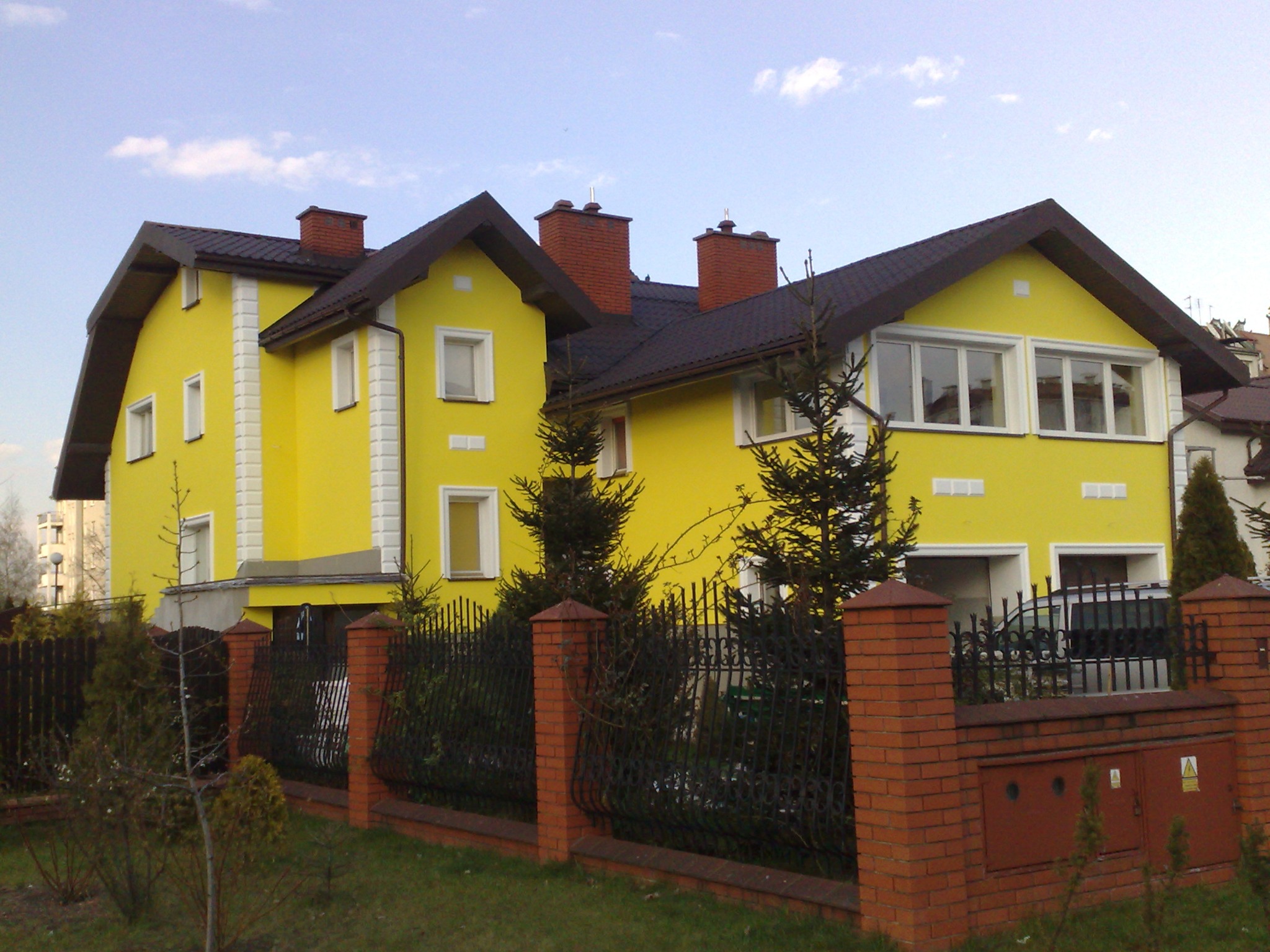 Żółty dom jednorodzinny z brązowym dachem i ceglanymi kominami, ogrodzony metalowym płotem z ceglanymi słupkami, widok z zewnątrz w słoneczny dzień.