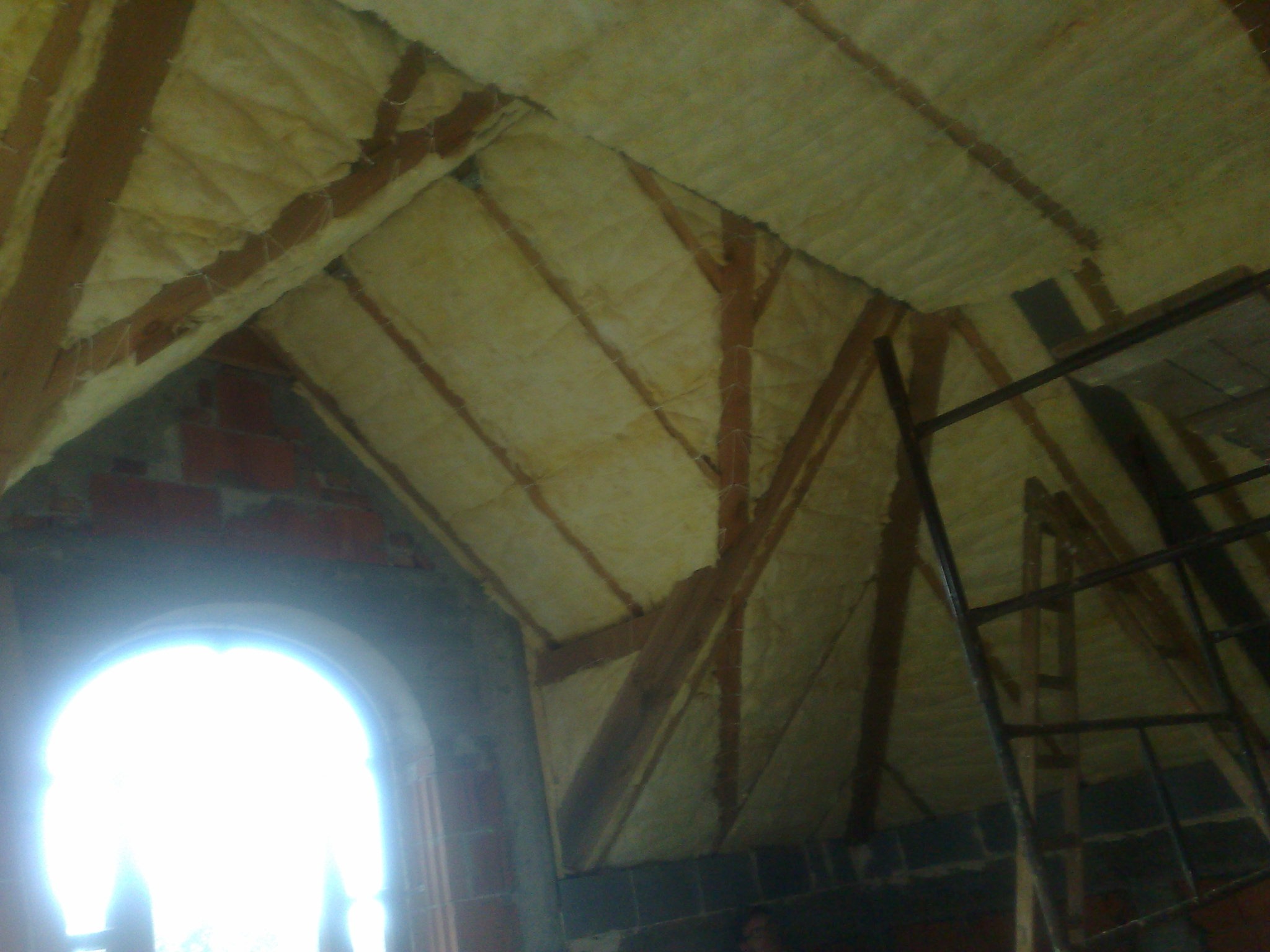 Izolacja poddasza wełną mineralną między krokwiami, widoczne fragmenty ceglanego muru i okno z intensywnym światłem oraz fragment rusztowania.