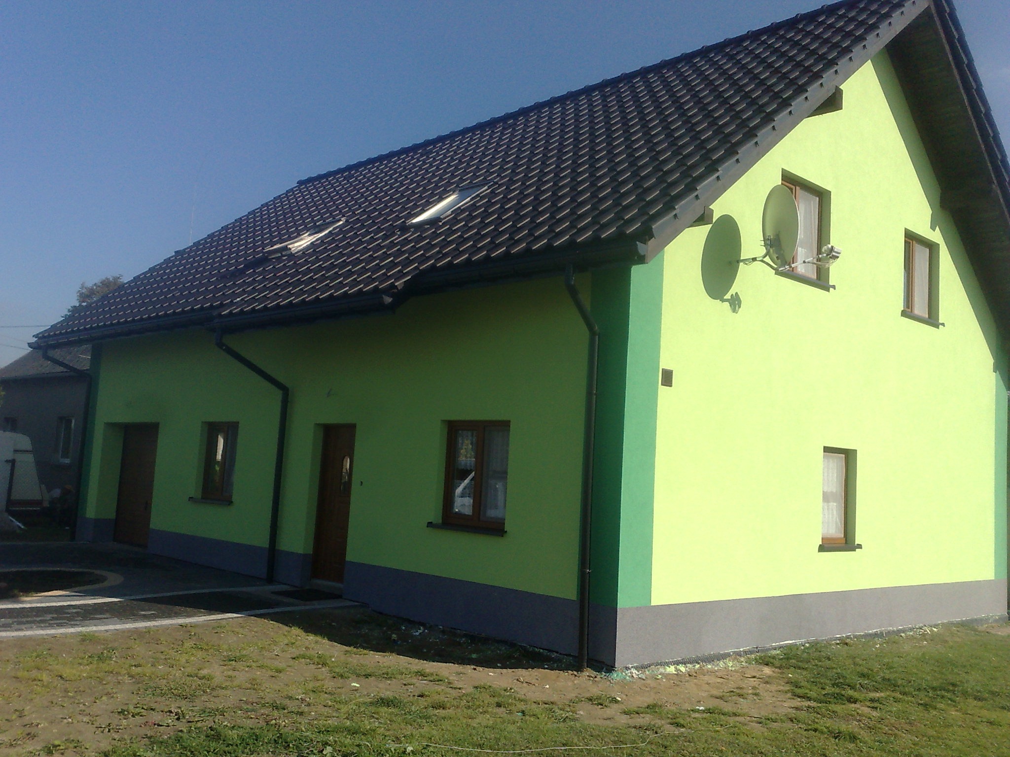 Nowo wybudowany dom jednorodzinny z zieloną elewacją i ciemną dachówką, widoczne dwie anteny satelitarne na ścianie szczytowej.