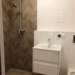 Łazienka w trakcie remontu: widoczny prysznic z deszczownicą na ścianie wyłożonej płytkami w jodełkę, biała umywalka z szafką, białe ściany i przygotowane podłoże pod podłogę.