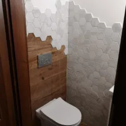 Nowoczesna toaleta we Wrocławiu z drewnianą boazerią do połowy ściany i białymi heksagonalnymi płytkami marmuropodobnymi ułożonymi schodkowo powyżej, widoczny podtynkowy stelaż WC z chromowanymi...