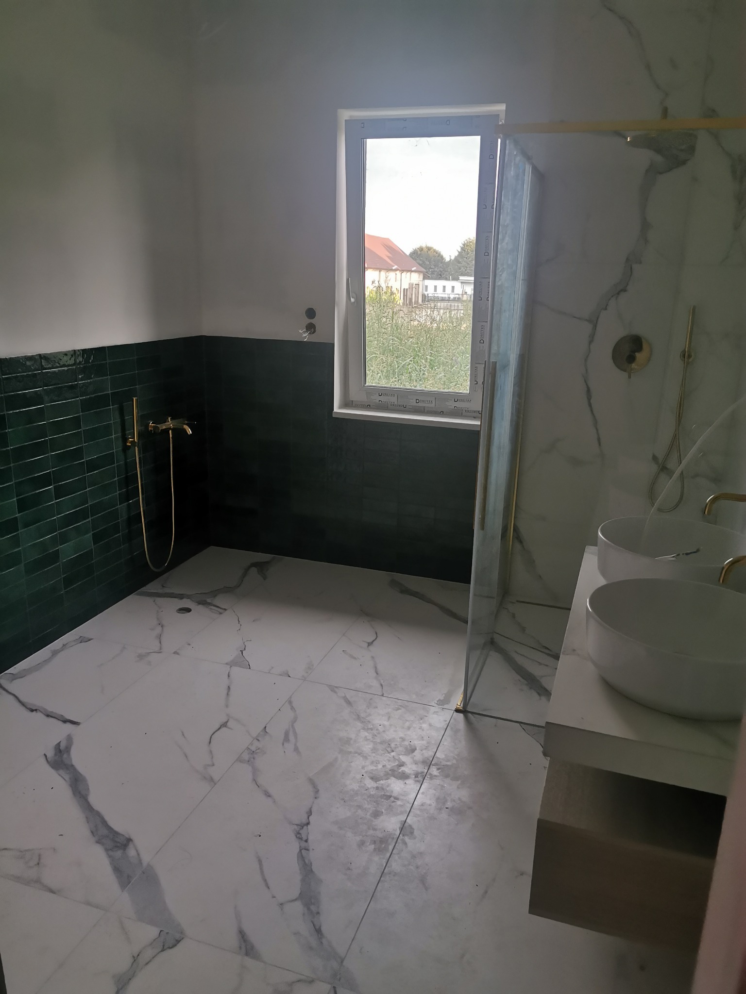 Nowoczesna łazienka z ciemnozielonymi płytkami na ścianie, marmurową podłogą i dwoma umywalkami na blacie, widok na zewnątrz przez okno