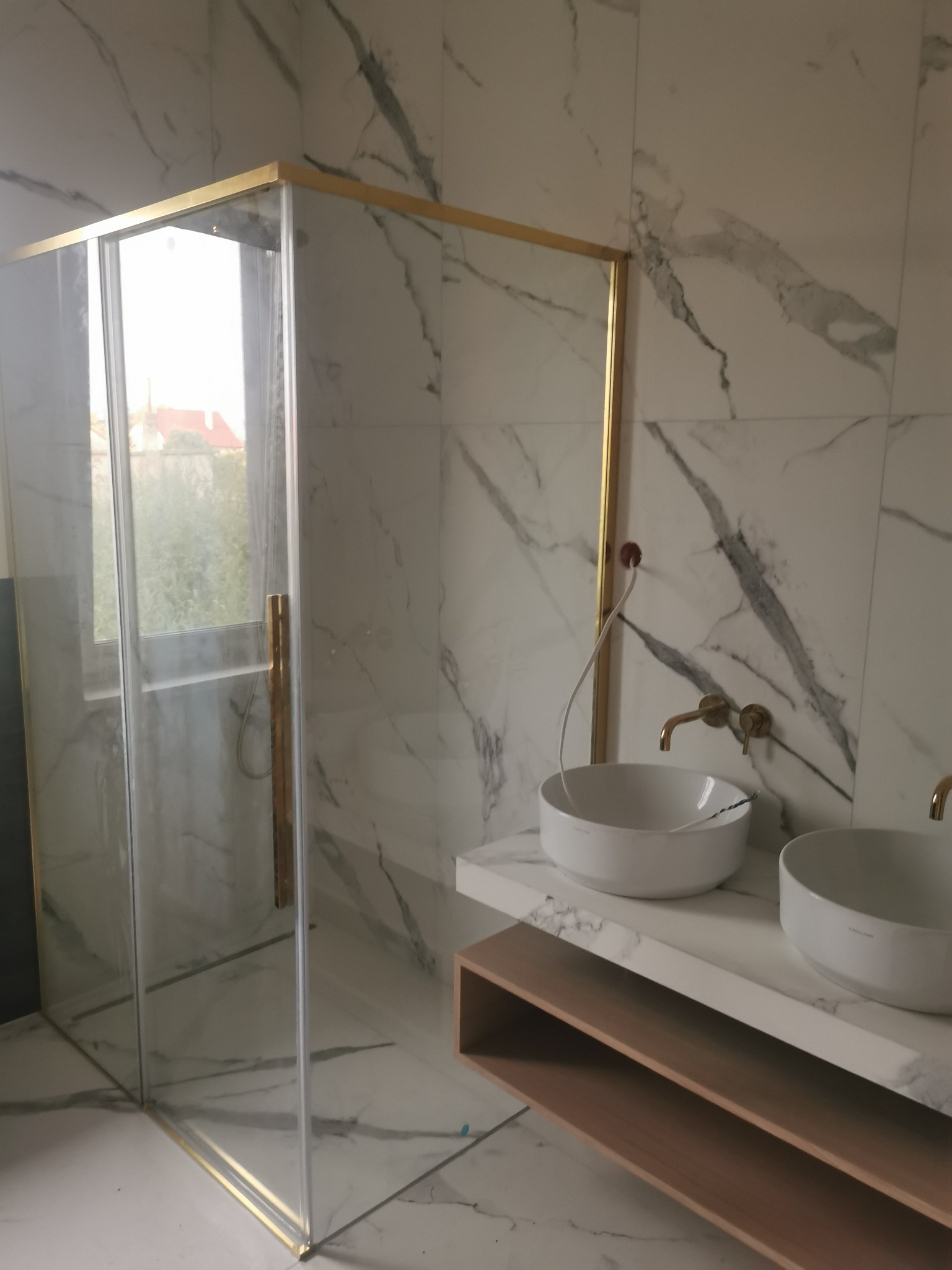 Nowoczesna łazienka z kabiną prysznicową z złotymi profilami, dwoma umywalkami nablatowymi na blacie z marmurowym wzorem oraz płytkami ściennymi i podłogowymi imitującymi marmur.