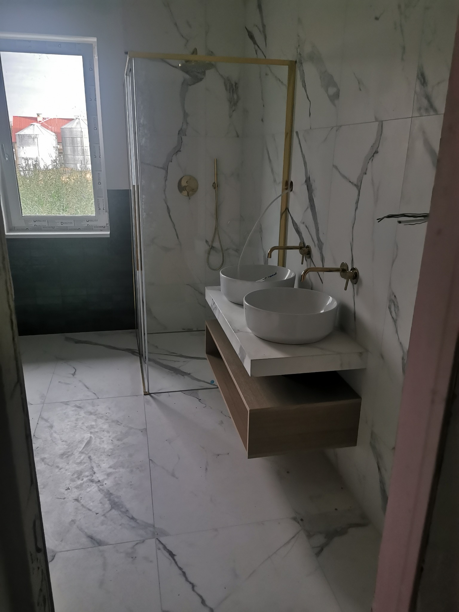Nowoczesna łazienka z marmurowymi płytkami, dwoma białymi umywalkami i złotą armaturą, widok na zewnątrz przez okno.