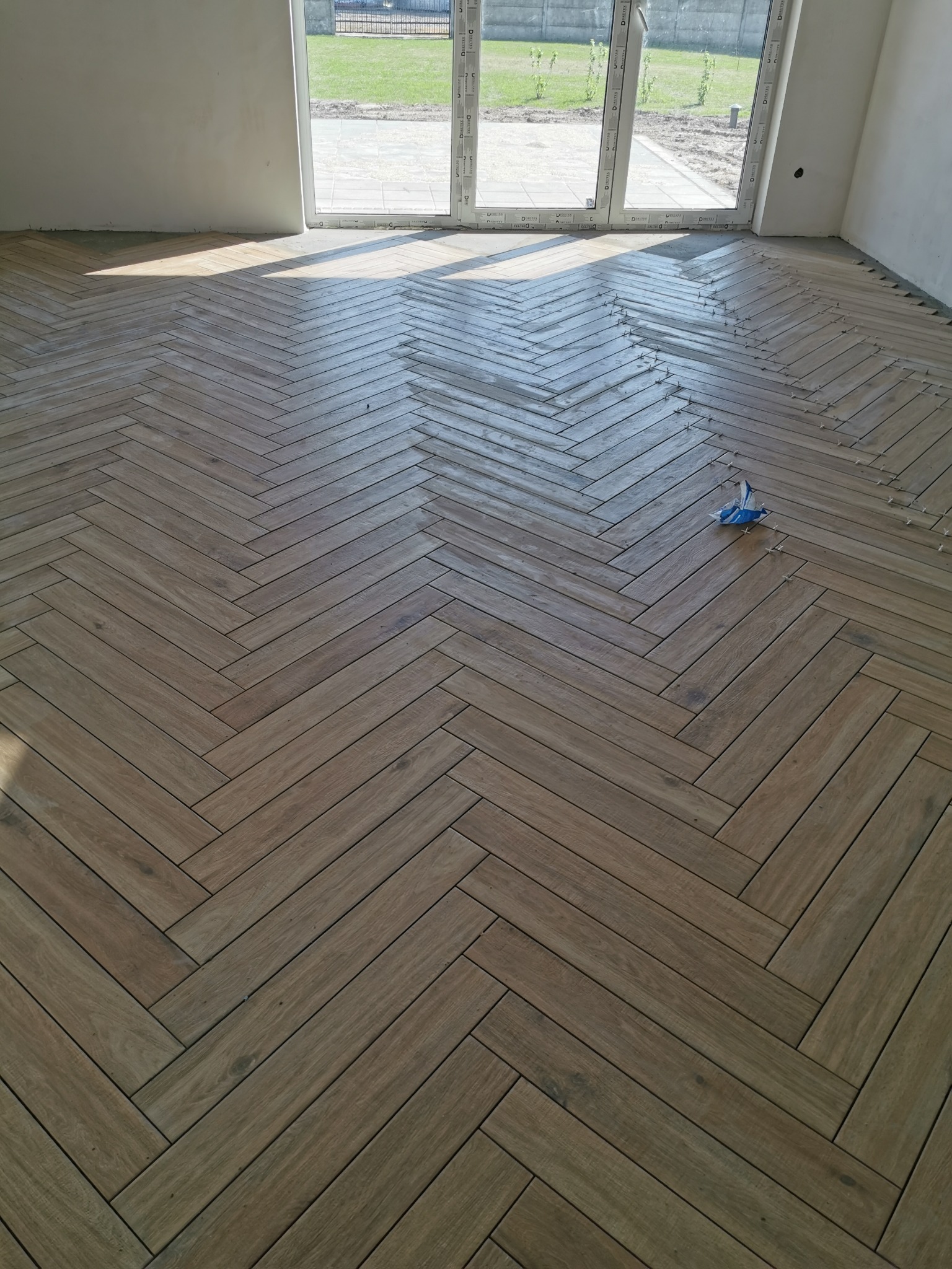 Układanie drewnopodobnych płytek w jodełkę francuską, widoczne krzyżyki dystansowe i resztki zaprawy, w tle okno z widokiem na ogród.