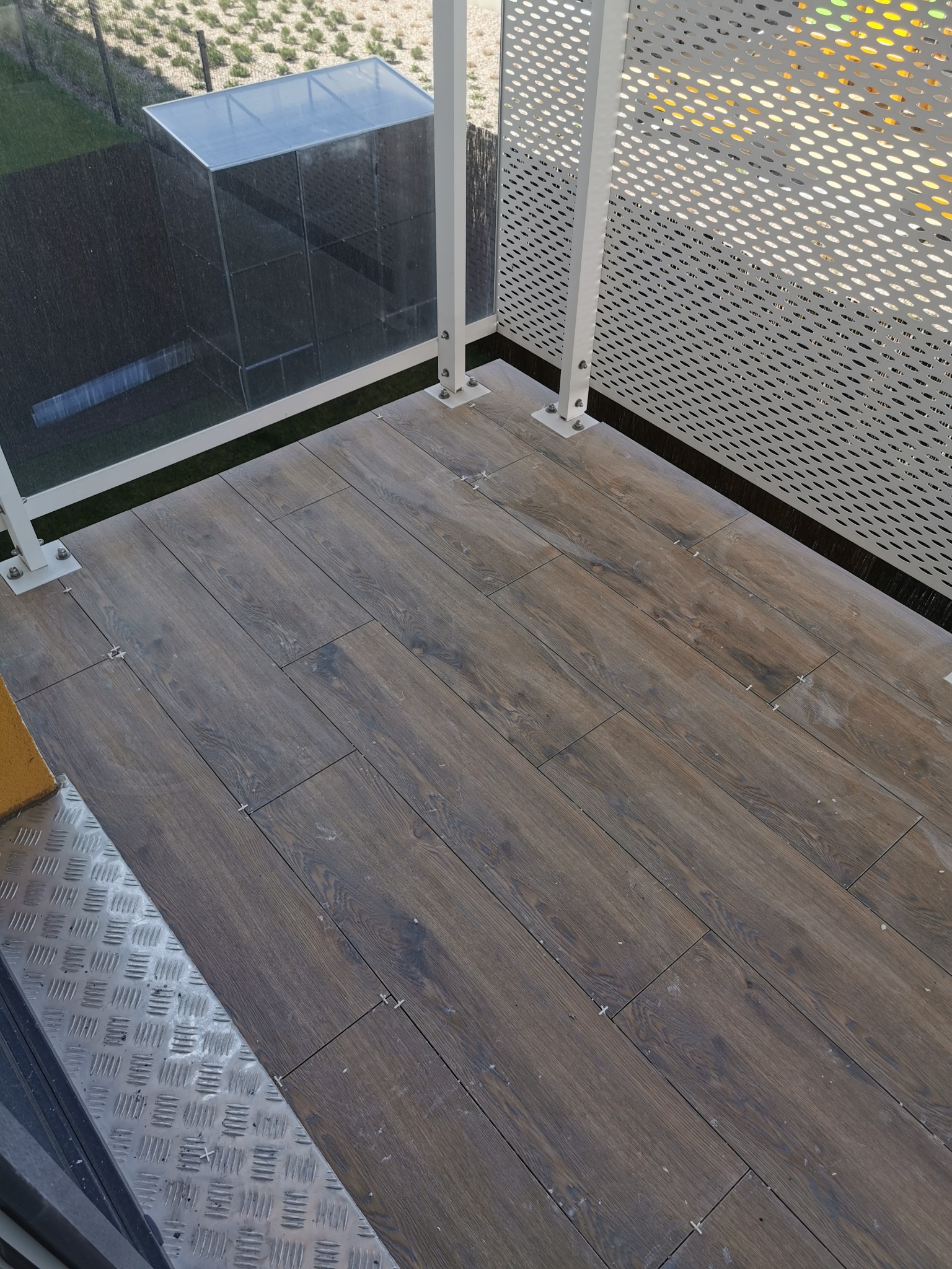 Balkon z nową, ciemnobrązową podłogą imitującą drewno, ułożoną z płytek ceramicznych z krzyżykami dystansowymi, biała ażurowa balustrada.