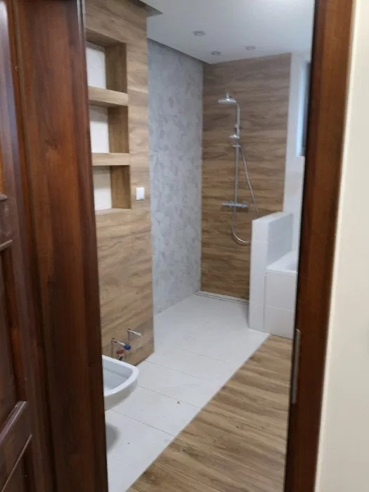 Nowoczesna łazienka z drewnopodobnymi płytkami, białą armaturą i kabiną prysznicową, widok zza drewnianych drzwi.