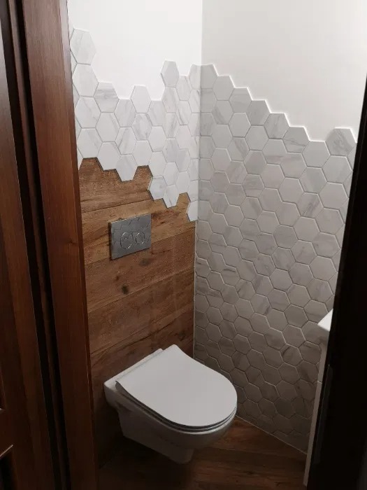 Nowoczesna toaleta we Wrocławiu z drewnianą boazerią do połowy ściany i białymi heksagonalnymi płytkami marmuropodobnymi ułożonymi schodkowo powyżej, widoczny podtynkowy stelaż WC z chromowanymi...