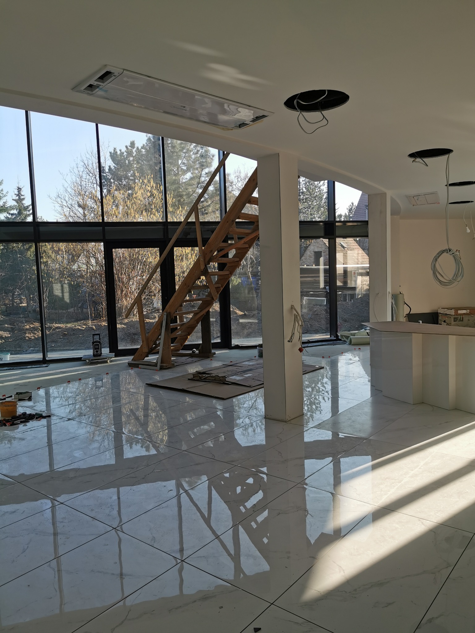 Wnętrze w trakcie prac wykończeniowych: duże okno z widokiem na ogród, drewniane schody tymczasowe, błyszcząca podłoga z płytek, widoczne narzędzia i materiały budowlane, przygotowane otwory...