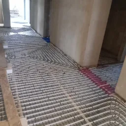 Białe rurki ogrzewania podłogowego ułożone na siatce montażowej w surowym wnętrzu budynku, widok z perspektywy podłogi.