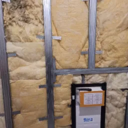 Instalacja stelaża podtynkowego WC marki Roca z izolacją z wełny mineralnej w Połczynie-Zdroju, widoczne metalowe profile konstrukcyjne i fragment nożyka monterskiego na obudowie.