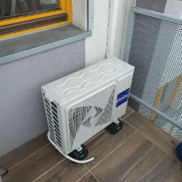 Biała jednostka zewnętrzna klimatyzatora Haier umieszczona na czarnych wspornikach z poziomnicami, zamontowana na balkonie z brązowymi płytkami imitującymi drewno, widoczna szara balustrada...