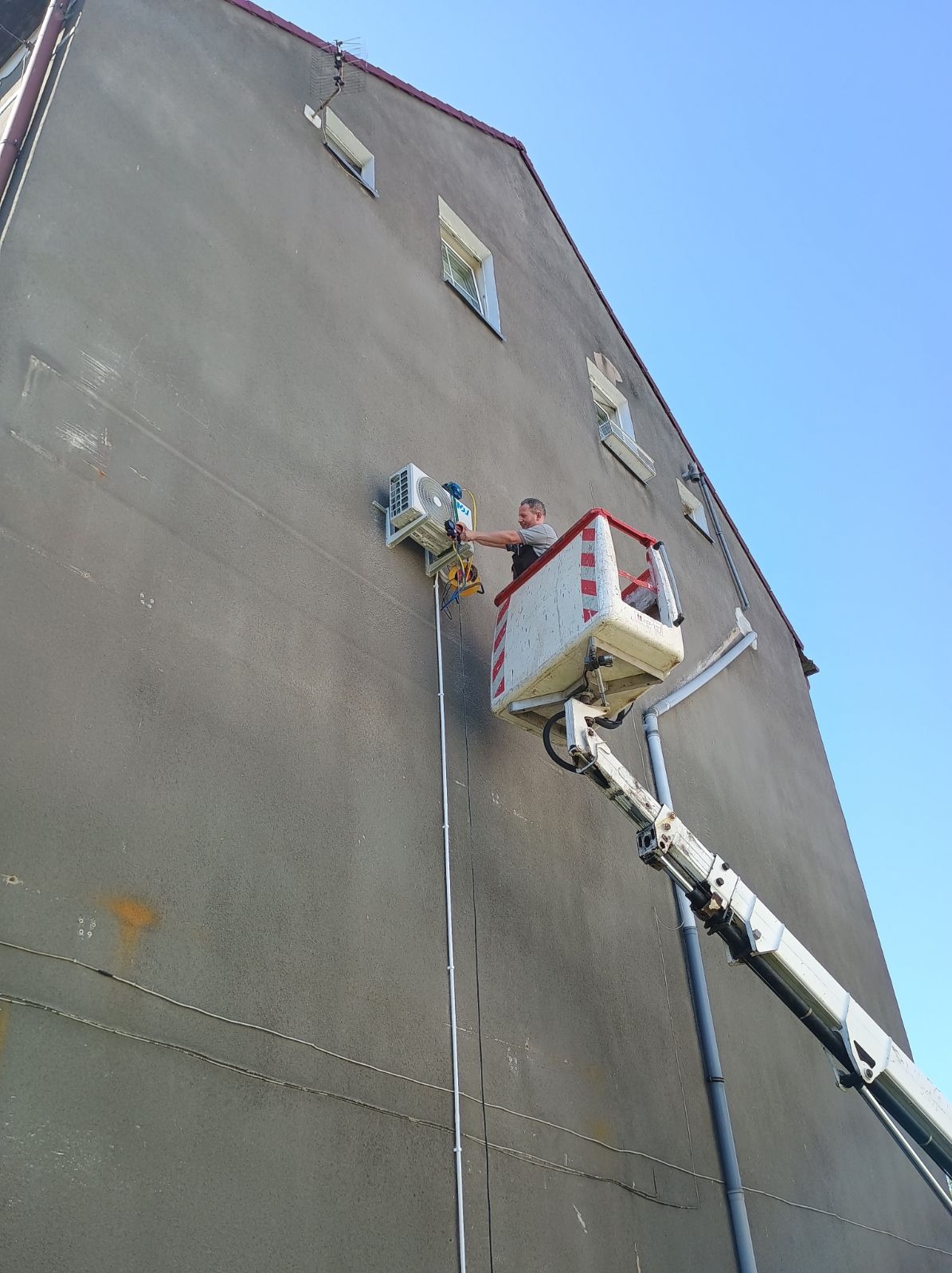 Pracownik na podnośniku koszowym montuje zewnętrzną jednostkę klimatyzacji na szarej ścianie budynku. Widoczne okna i instalacje na elewacji.