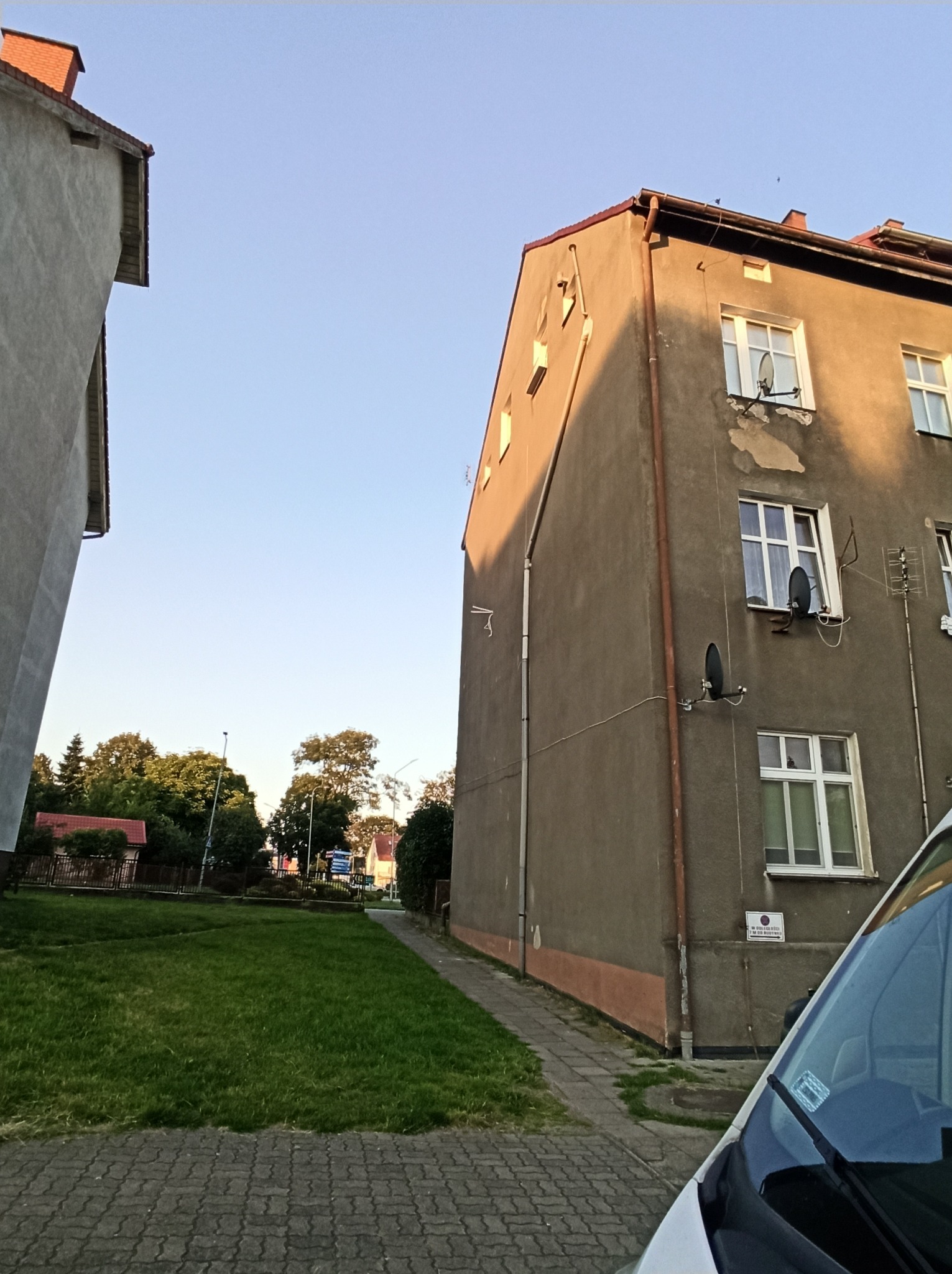 Fragment elewacji bloku mieszkalnego z widocznymi antenami satelitarnymi, rurami spustowymi i tabliczką informacyjną, sfotografowany w słoneczny dzień.