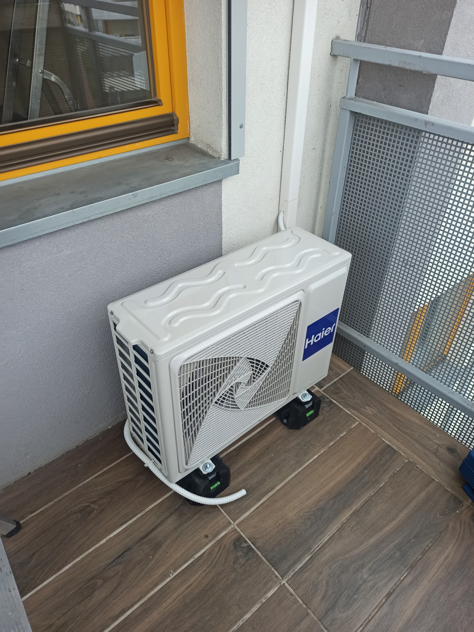 Biała jednostka zewnętrzna klimatyzatora Haier zamontowana na czarnych podkładkach antywibracyjnych na balkonie z drewnopodobnymi płytkami, widoczna rura odprowadzająca skropliny.