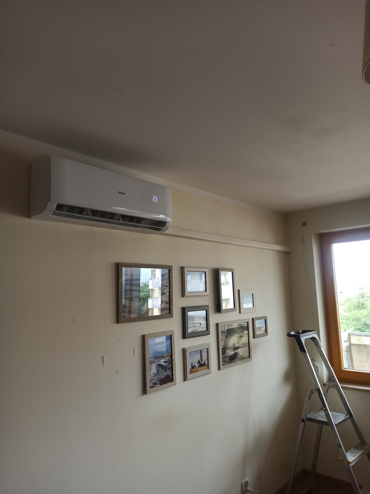Biały klimatyzator Haier zamontowany na ścianie w pokoju, pod nim galeria obrazków w ramkach, obok okna rozstawiona aluminiowa drabina.