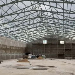 Wnętrze hali w trakcie budowy z widoczną konstrukcją stalową dachu, betonowymi ścianami i surową posadzką. Widoczne okna i materiały budowlane.