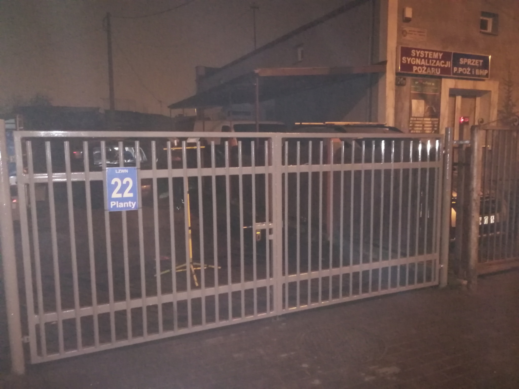 Szara, metalowa brama z pionowymi prętami i niebieską tabliczką z numerem '22' przy wejściu do posesji, widoczna w nocy, oświetlona ulicznymi lampami.