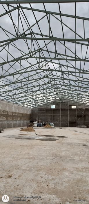 Wnętrze hali w trakcie budowy z widoczną konstrukcją stalową dachu, betonowymi ścianami i surową posadzką. Widoczne okna i materiały budowlane.