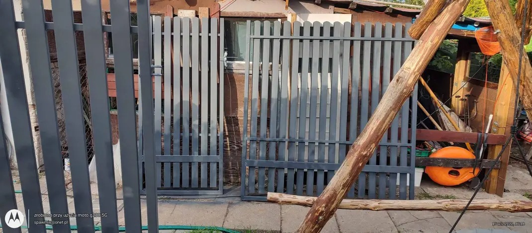 Szara, metalowa brama z pionowymi szczeblami w trakcie montażu, widoczne elementy konstrukcyjne i tło z fragmentami budynku.