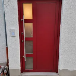 Czerwone drzwi zewnętrzne z trzema pionowymi szybami i długim, srebrnym uchwytem, zamontowane w białej, teksturowanej ścianie z domofonem.