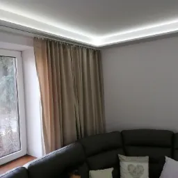 Nowoczesne wnętrze z oknem, zasłonami w kolorze beżowym i podświetleniem LED na suficie, widok na zimowy ogród.