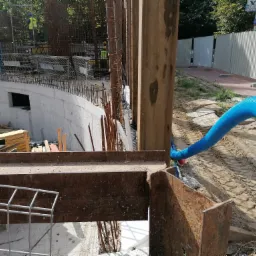 Budowa podziemnej kondygnacji budynku z widocznymi stalowymi elementami konstrukcyjnymi, szalunkiem, zbrojeniami oraz niebieskim wężem wentylacyjnym.