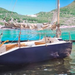 TOM-BUD Zbigniew Tomaszewski - Scena filmowa z jachtem na wodzie przed ekranem imitującym krajobraz morski; mężczyzna w hawajskiej koszuli czyta książkę na pomoście, a drugi, w woderach, stoi w wodzie z kijem.