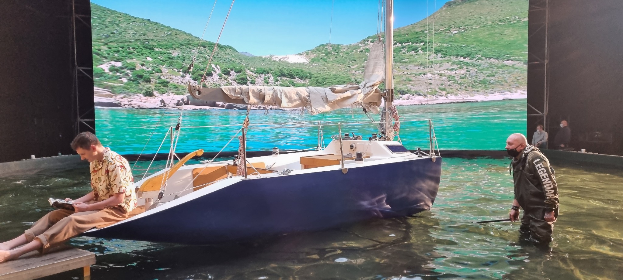 Scena filmowa z jachtem na wodzie przed ekranem imitującym krajobraz morski; mężczyzna w hawajskiej koszuli czyta książkę na pomoście, a drugi, w woderach, stoi w wodzie z kijem.