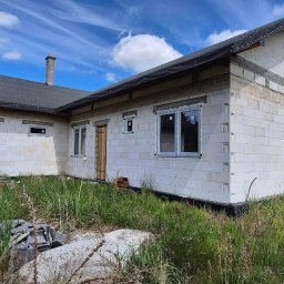 MB OKNA I DRZWI Dariusz Nowakowski - Surowy stan domu z białego pustaka z zamontowanymi oknami PCV, widok z zewnątrz, wysoka trawa na pierwszym planie, błękitne niebo z chmurami w tle.