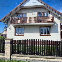 MB OKNA I DRZWI Dariusz Nowakowski - Dom jednorodzinny z brązowymi oknami PCV i balkonem z drewnianą balustradą, otoczony czarnym płotem z kamienną podmurówką, w słoneczny dzień.