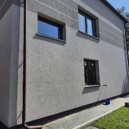 Szary dom z trzema oknami o ciemnych ramach i brązową rynną. Elewacja z poziomymi pasami w odcieniach szarości. Ujęcie z poziomu chodnika.