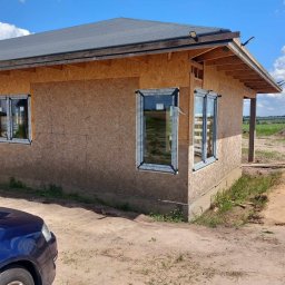 MB OKNA I DRZWI Dariusz Nowakowski - Dom w trakcie budowy, z zamontowanymi białymi oknami w ścianach z płyty OSB. W tle widoczne pola i niebo.