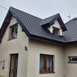 MB OKNA I DRZWI Dariusz Nowakowski - Dom z beżową elewacją i brązowymi oknami PCV, czarny dach z dachówki, widok z dołu, pochmurne niebo. Zewnętrzna lampa ścienna.