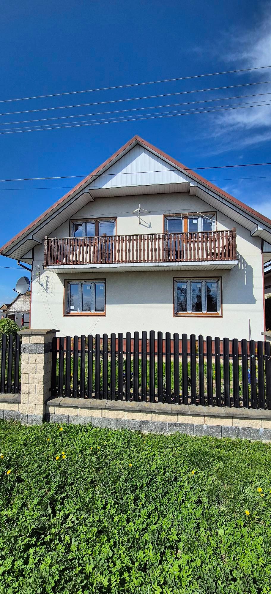 Dom jednorodzinny z brązowymi oknami PCV i balkonem z drewnianą balustradą, otoczony czarnym płotem z kamienną podmurówką, w słoneczny dzień.