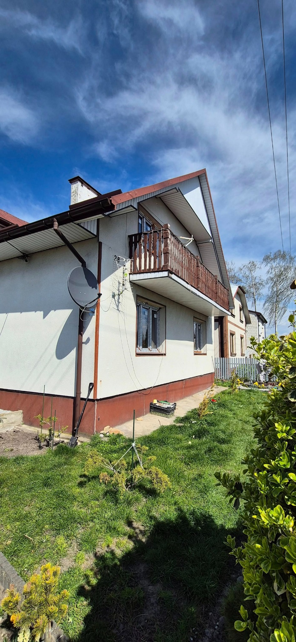 Dwupiętrowy dom z balkonem z drewnianą balustradą, okna z brązowymi ramami, antena satelitarna na elewacji. Błękitne niebo w tle, trawnik na pierwszym planie.