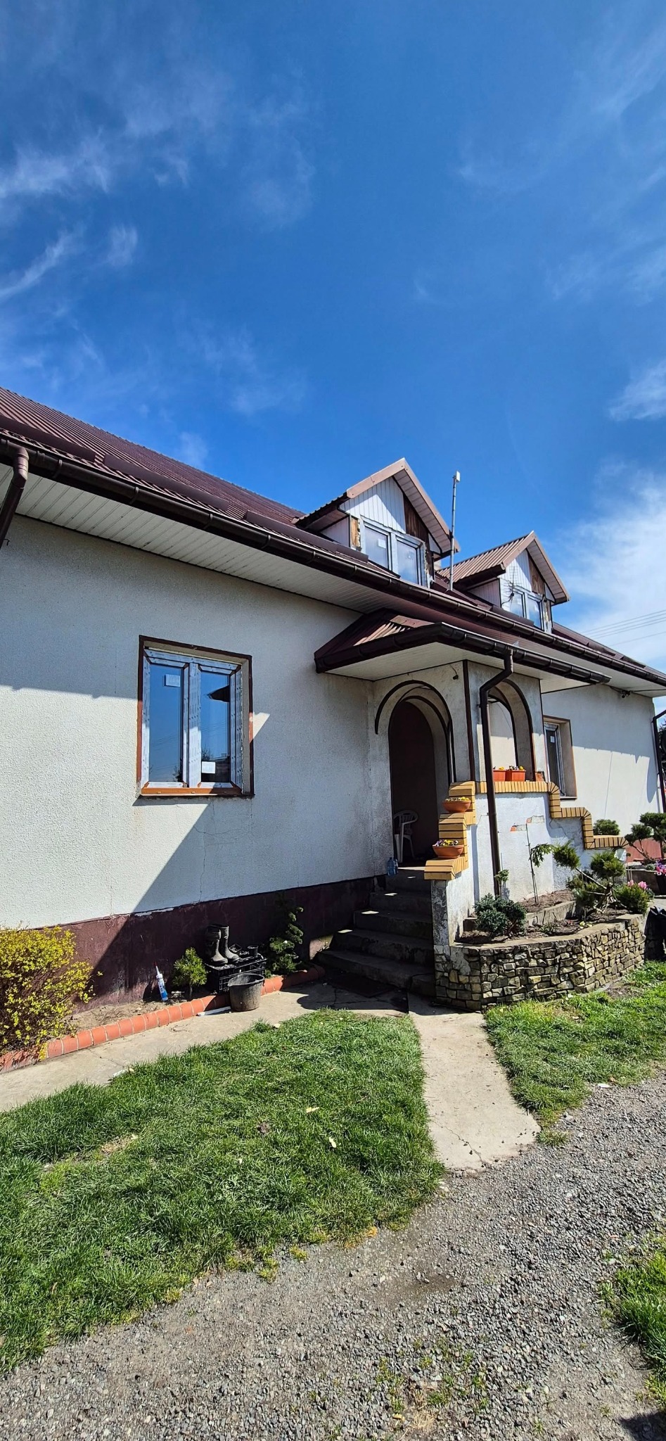 Dom jednorodzinny z oknami w trakcie montażu, widoczne niezabezpieczone ramy okienne. Częściowo otynkowana elewacja i brązowy dach. Błękitne niebo w tle.