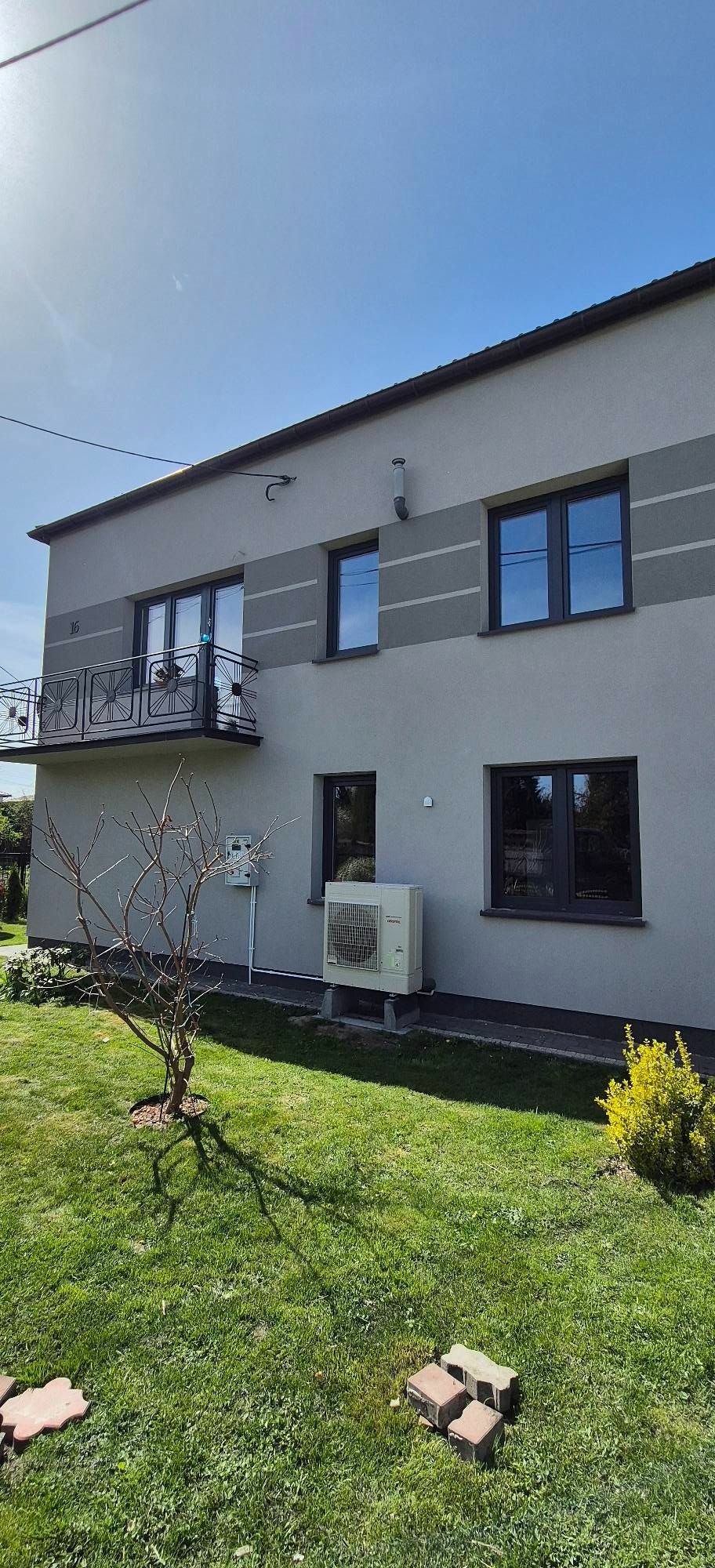 Elewacja domu z ciemnymi oknami, balkonem z ozdobną balustradą i klimatyzatorem na zewnątrz. Widok na zadbany trawnik i błękitne niebo.