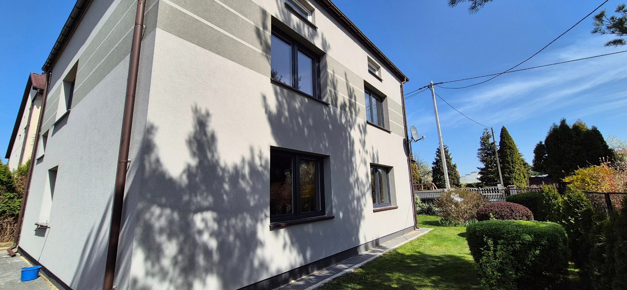 Elewacja domu z ciemnymi oknami, widok z zewnątrz, w tle błękitne niebo i zieleń ogrodu. Na ścianie cień drzewa. Widoczny fragment rynny i satelity.