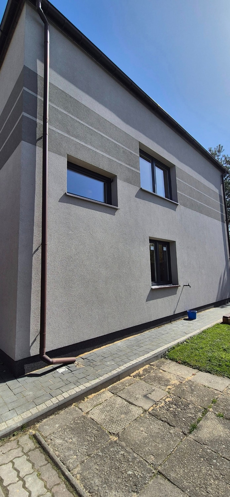 Szary dom z trzema oknami o ciemnych ramach i brązową rynną. Elewacja z poziomymi pasami w odcieniach szarości. Ujęcie z poziomu chodnika.