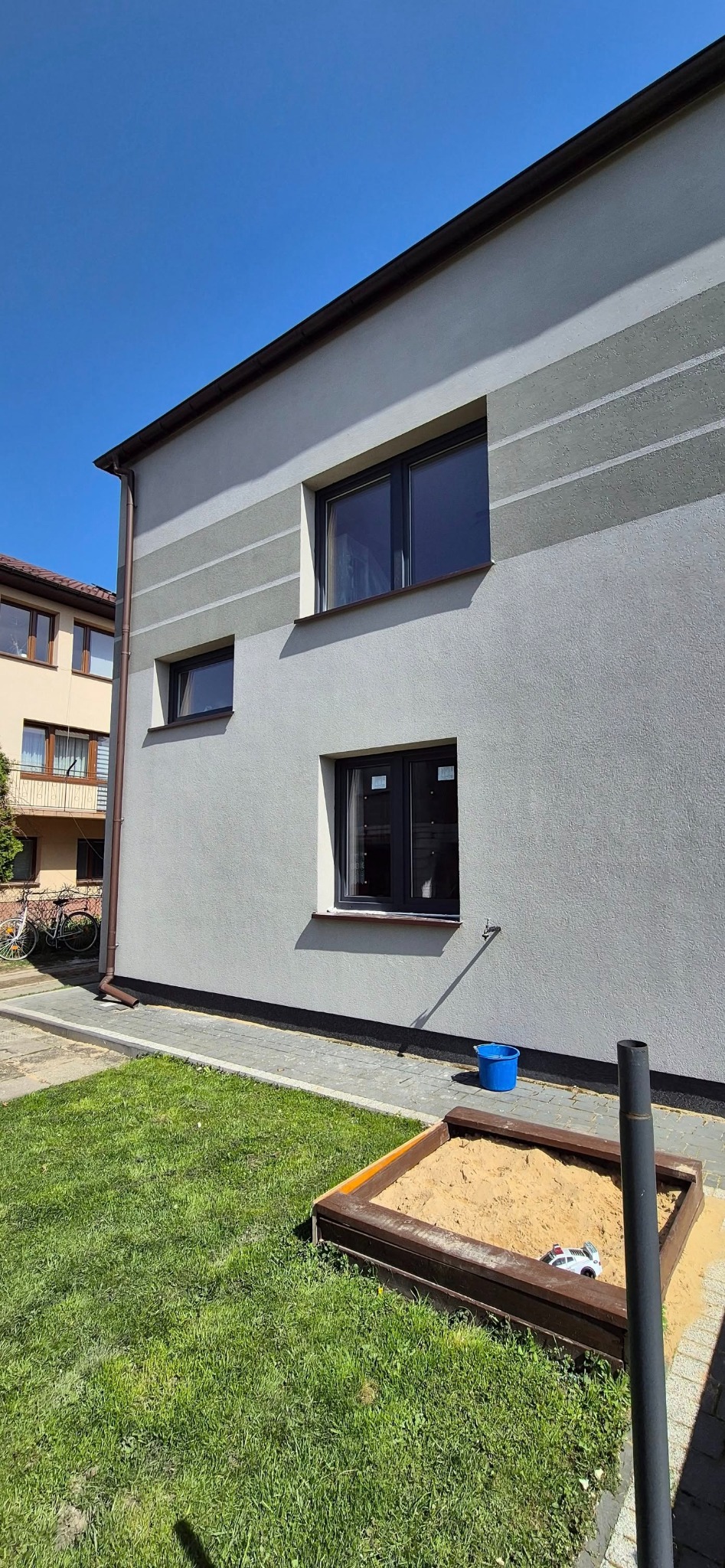 Elewacja domu z trzema oknami w ciemnych ramach, widok z boku. Na pierwszym planie trawnik i piaskownica z zabawką, błękitne niebo w tle.